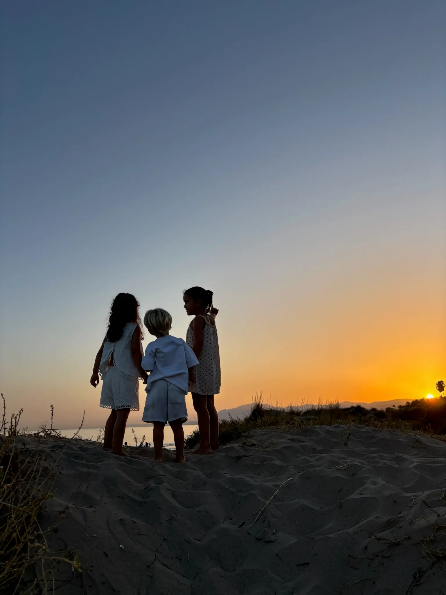 Tres niños en una duna al atardecer, mirando el mar.